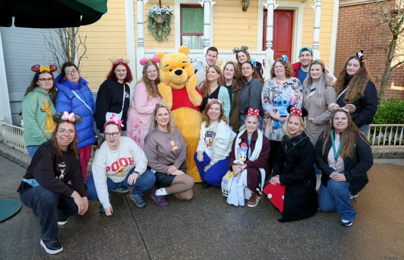 Een grote groep Pixies staat en zit rond Winnie de Poeh voor een geel huisje in Disneyland Parijs